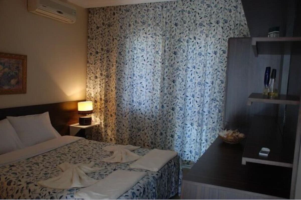 A hotel room with a bed, a nightstand with a lamp, a window with floral curtains, and a shelf with toiletries.