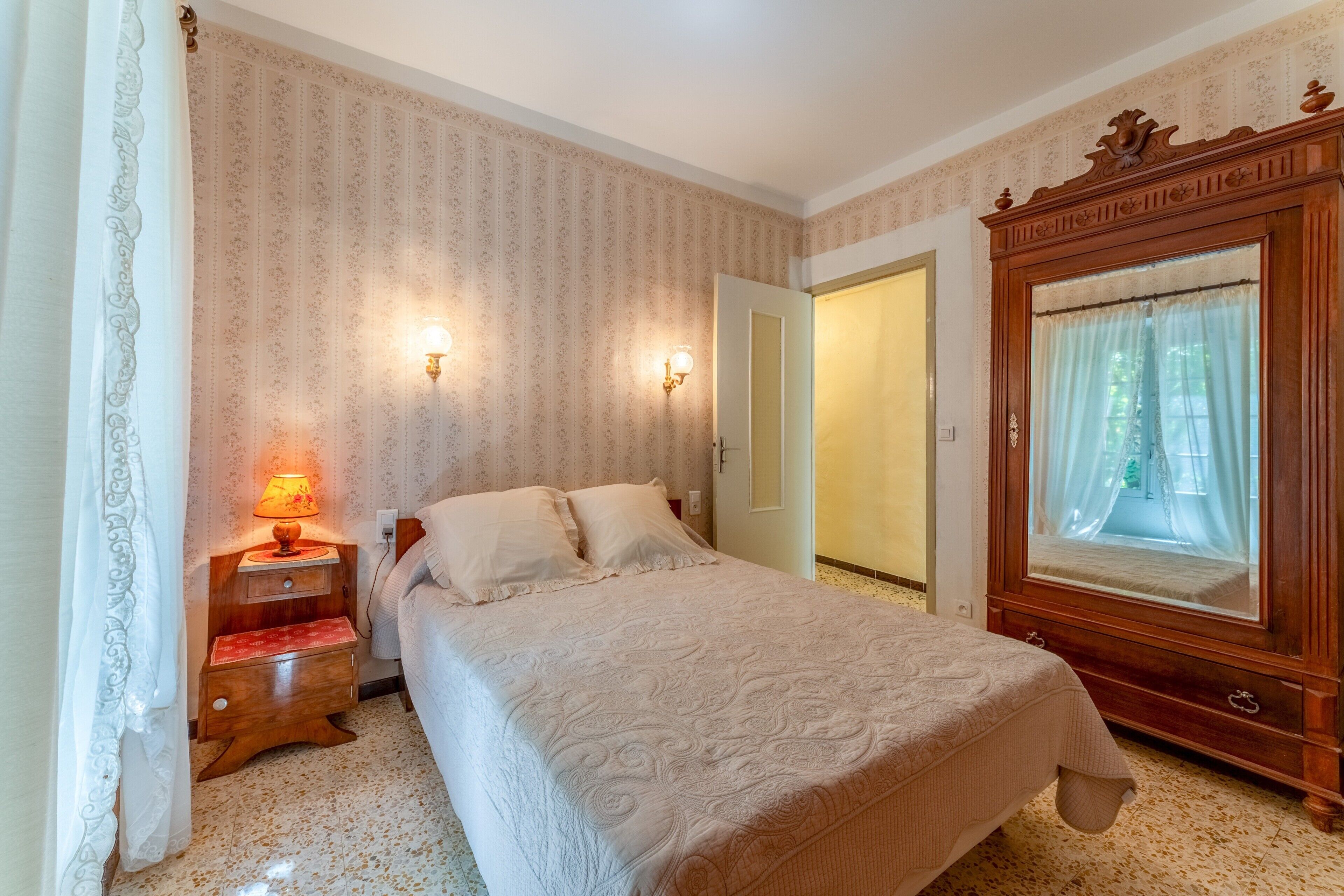 A bedroom with a bed, a nightstand with a lamp, a mirror, and patterned wallpaper.
