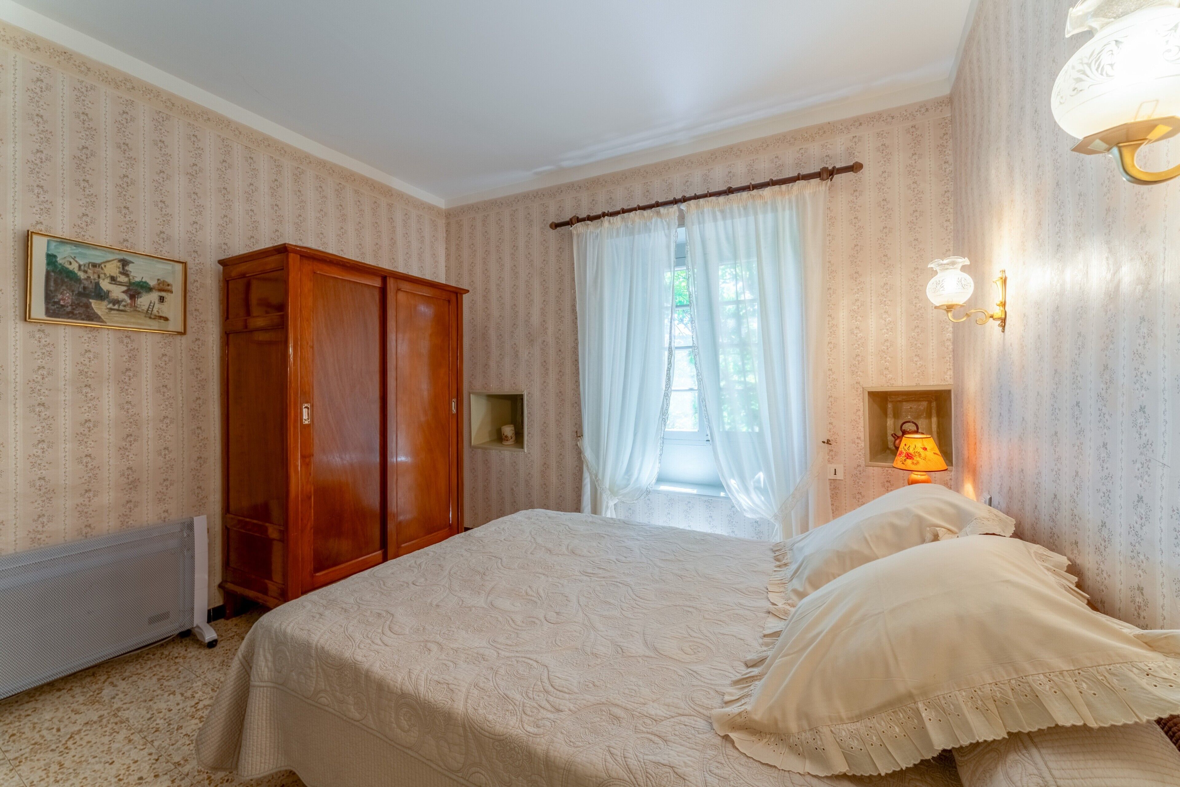 Una camera da letto con un letto, un armadio, un termosifone, una finestra con tende e una lampada a parete.