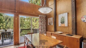 A wooden dining area with a table and chairs, a hanging light fixture, and a view of a forested area through a glass door.