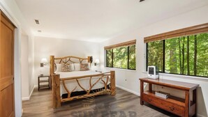 Un dormitorio con una cama grande, cabecera de madera, dos mesitas de noche, un tocador y un aparador de madera, además de grandes ventanas con vistas a un bosque.
