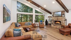 Una amplia sala de estar con un sofá modular grande, una chimenea y ventanas grandes que ofrecen vistas a los árboles.
