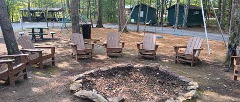 A campsite with a fire pit surrounded by Adirondack chairs, a swing set, and a small cabin in the background.