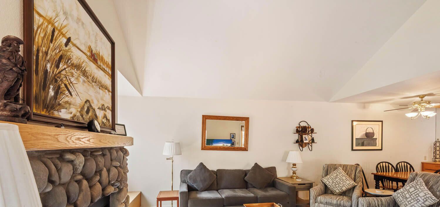 A living room with a stone fireplace, a grey sofa, a patterned armchair, a wooden table, and a ceiling fan.