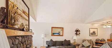 A living room with a stone fireplace, a grey sofa, a patterned armchair, a wooden table, and a ceiling fan.