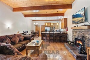 Uma sala de estar ampla com lareira, teto de madeira e cozinha ao fundo.