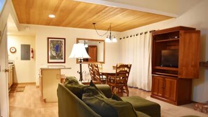 A cozy living room with a wooden ceiling, a green sofa, a dining table with chairs, a TV cabinet, and a lamp.