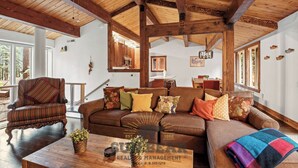 A spacious living room with a large wooden ceiling, a brown sofa set, a wooden coffee table, and a view of the outdoors through large windows.