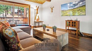 A living room with a wooden ceiling, a sofa with floral cushions, a coffee table, a lamp, a deer head decoration, a cabinet with glass doors, and a painting of a landscape.