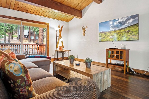 A living room with a wooden ceiling, a sofa with floral cushions, a coffee table, a lamp, a deer head decoration, a cabinet with glass doors, and a painting of a landscape. - 3 Bedroom Open Space Condo (IC3-72) (Incline Village)
