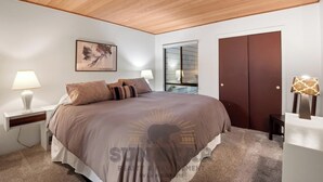 A bedroom with a large bed, two bedside tables with lamps, a wooden ceiling, a framed artwork, a sliding door, and a carpeted floor. - 3 Bedroom w Summer Pool and Hot Tub (FP1-83) (Incline Village)