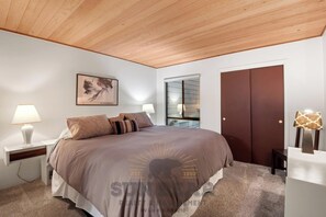 A bedroom with a large bed, two bedside tables with lamps, a wooden ceiling, a framed artwork, a sliding door, and a carpeted floor. - 3 Bedroom w Summer Pool and Hot Tub (FP1-83) (Incline Village)