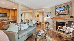 A living room with a fireplace, a television mounted on the wall, and a kitchen area with wooden cabinets.