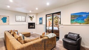 A spacious living room with a large sectional sofa, a black leather armchair, and a fireplace. There are several wall-mounted artworks, including a map, a bear painting, and a scenic landscape. - 3 Bedroom w Hot Tub and Forest Views (829EL) (Incline Village)