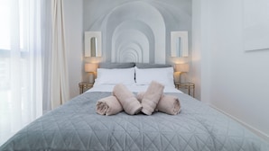 A neatly made bed with a quilted grey bedspread, two white pillows, and rolled towels on the bed. There are two bedside lamps on either side of the bed.