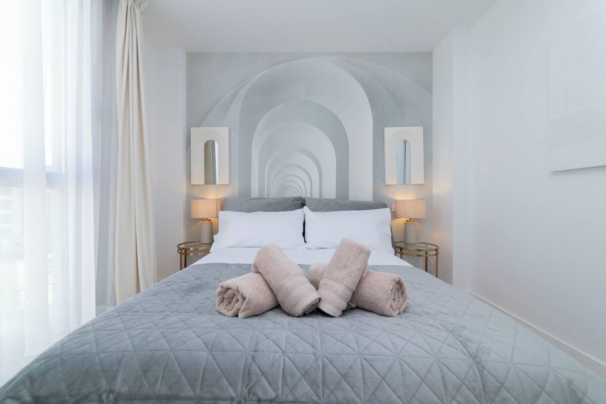 A neatly made bed with a quilted grey bedspread, two white pillows, and rolled towels on the bed. There are two bedside lamps on either side of the bed.
