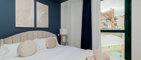 A hotel room with a bed, a nightstand, a lamp, and a view of buildings through a window.