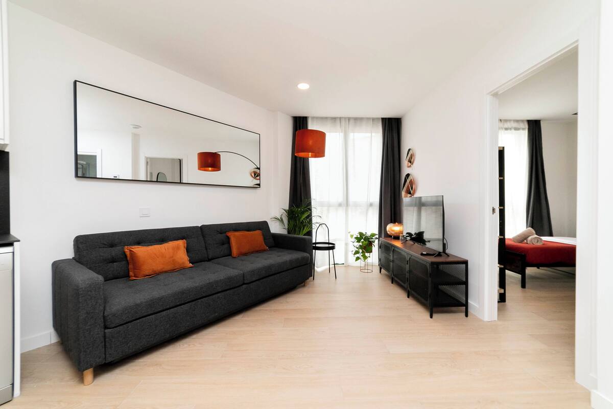 A modern living room with a grey sofa, a TV, and a red bed in the adjacent room.