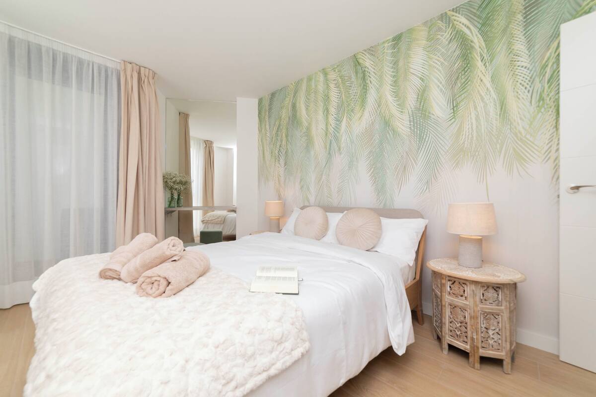 A neatly made bed with rolled towels, a bedside table with a lamp, and a wall with a palm leaf pattern.