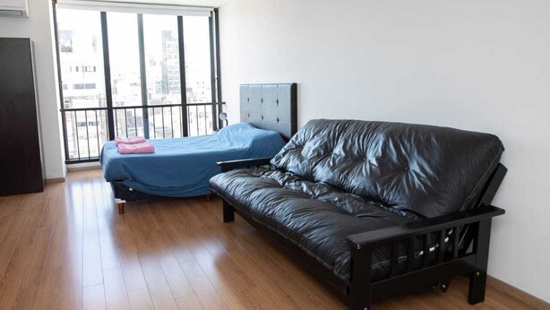 A room with a black leather sofa, a blue bed with a pink blanket, and a window with a view of buildings. - Rosario Center (Rosario)