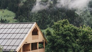 Una cabaña de madera con techo inclinado y ventanas amplias, rodeada de un bosque denso y montañas cubiertas de bruma.
