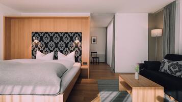 A modern hotel room with a large bed, a wooden headboard, a black and white patterned wall, a black sofa, a small table with a vase, and a view into the hallway.