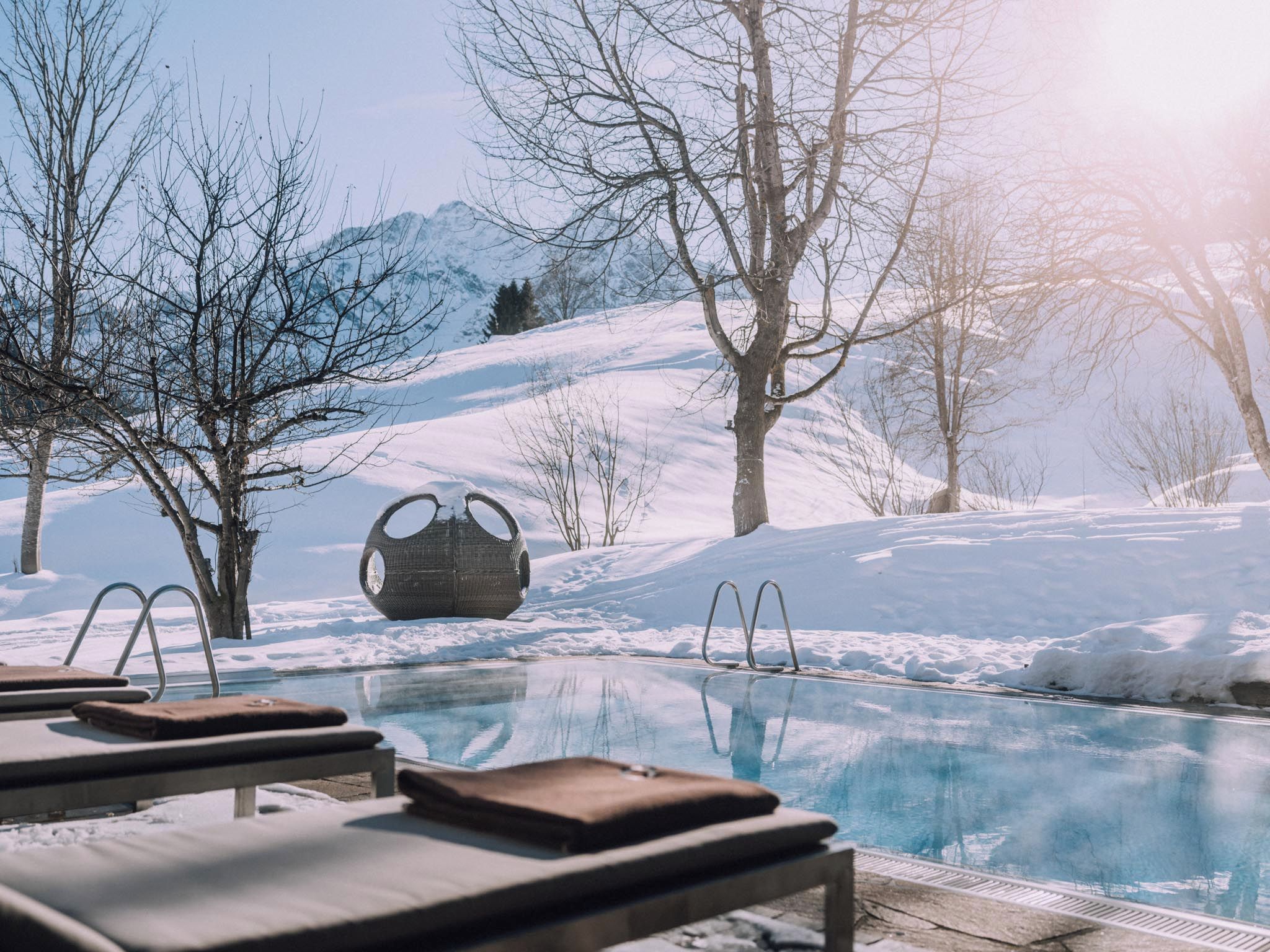 Un área de piscina con sillones de descanso y una original silla esférica, rodeada por un paisaje nevado y árboles.
