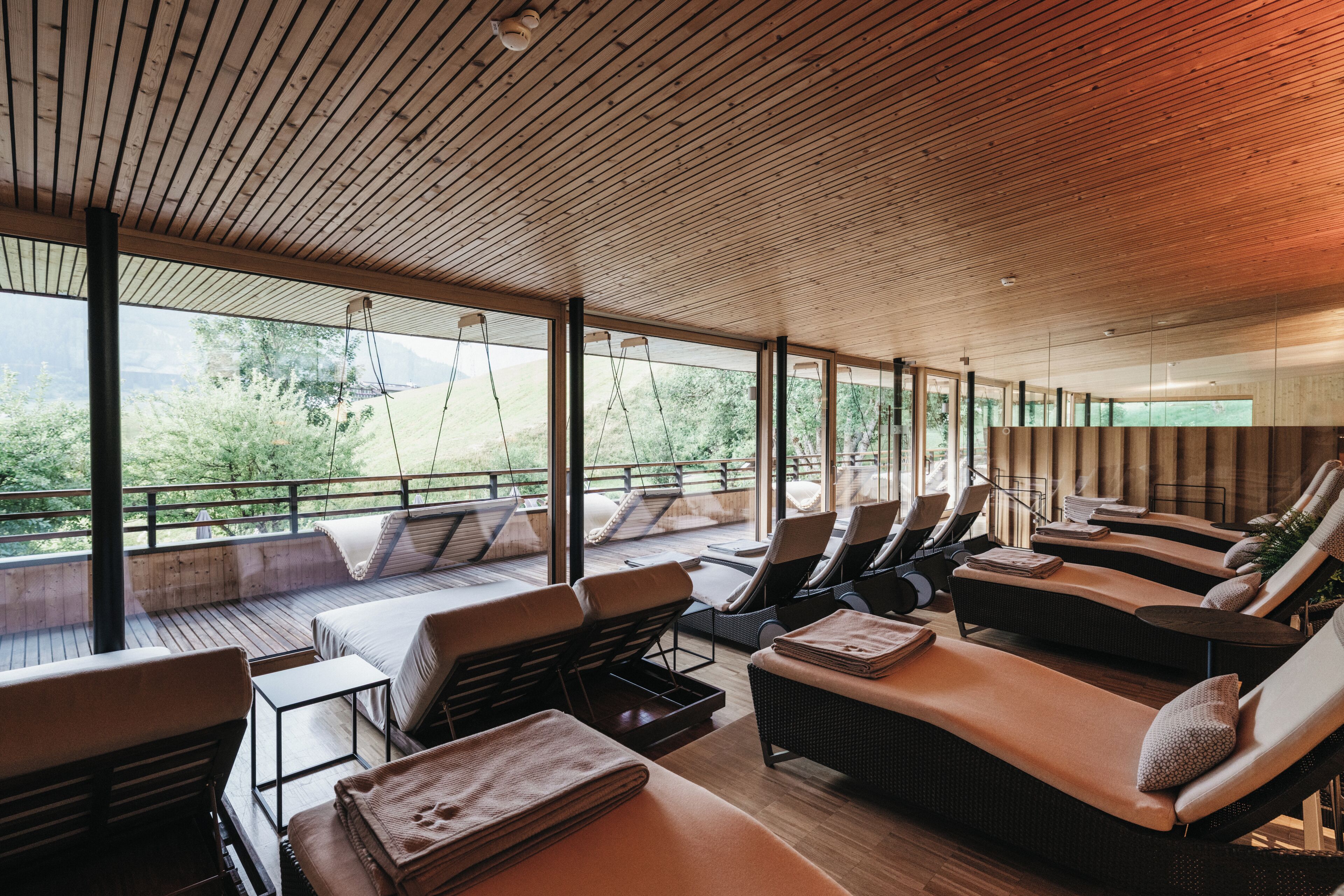 A modern outdoor lounge area with wooden flooring, cushioned seating, and large windows offering a view of greenery.