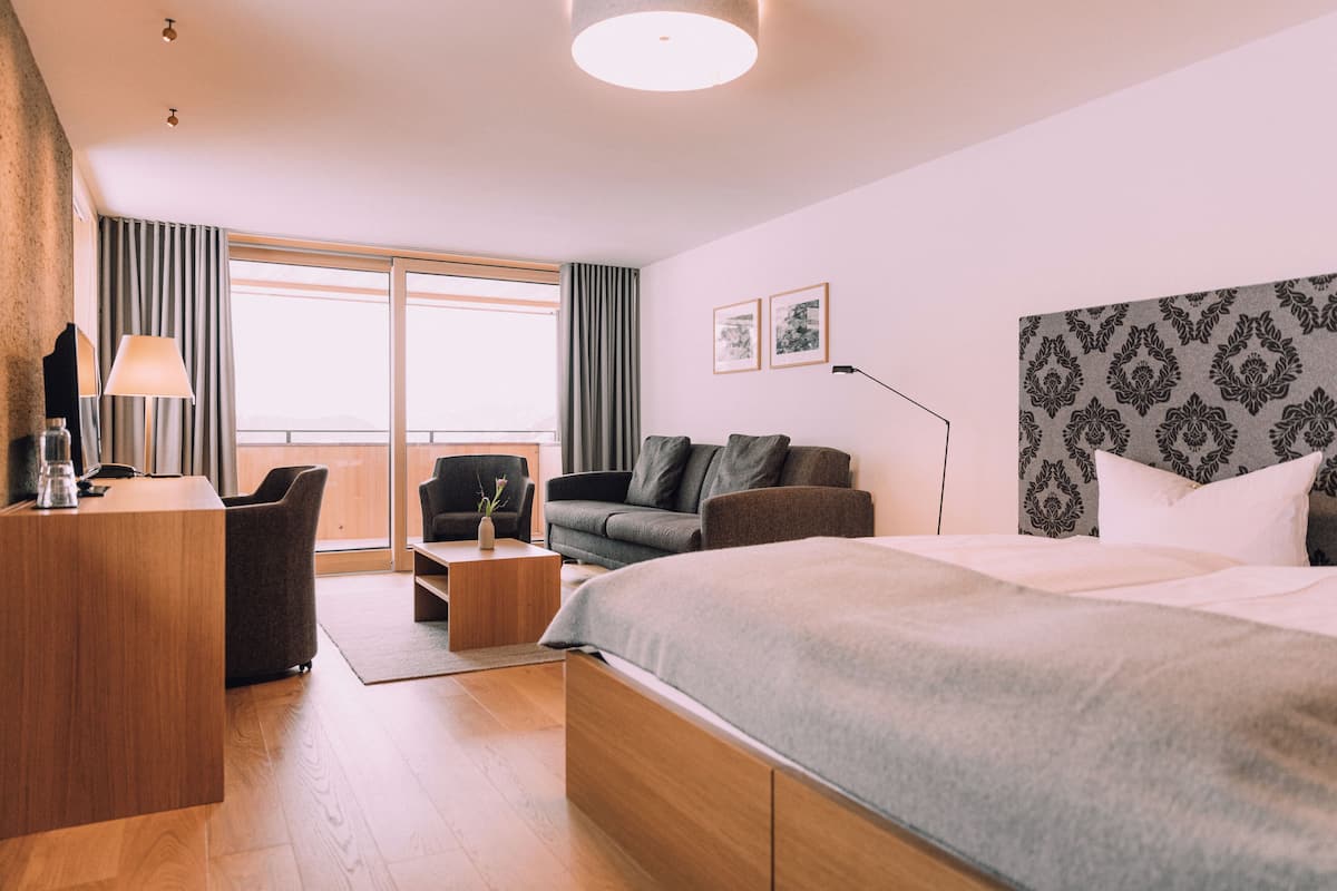 A modern hotel room with a large bed, a desk, a sofa, and a view of the outdoors.