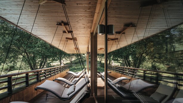 Terraza de madera con sillones reclinables y barandilla de vidrio, con vista a un área boscosa.