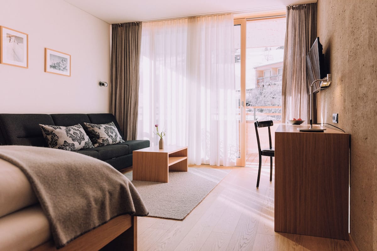 A modern hotel room with a bed, a desk, a chair, a small table, and a view of the city through large windows.