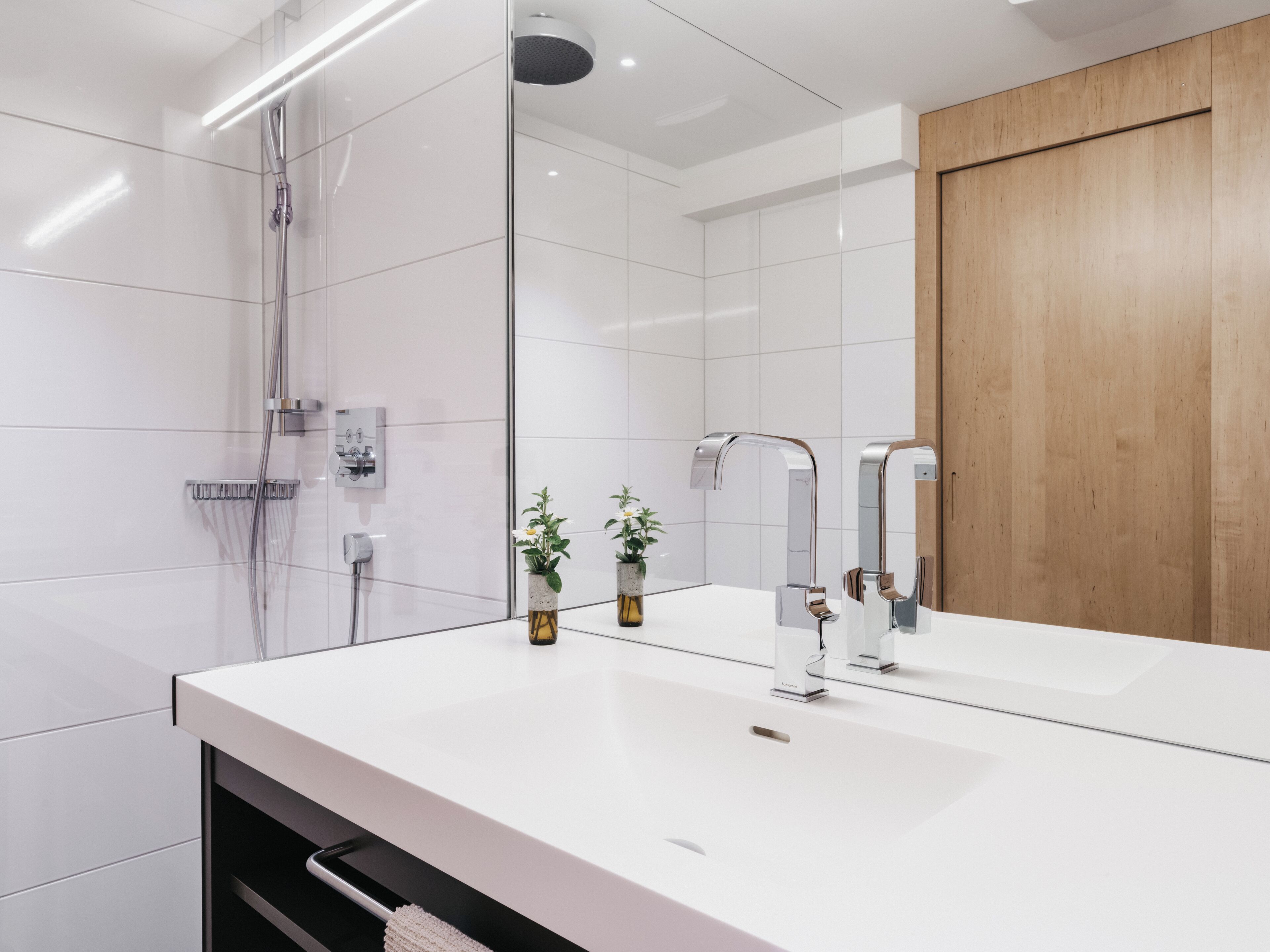 Un baño moderno con un espejo grande, baldosas blancas, una puerta de madera y una zona de ducha.