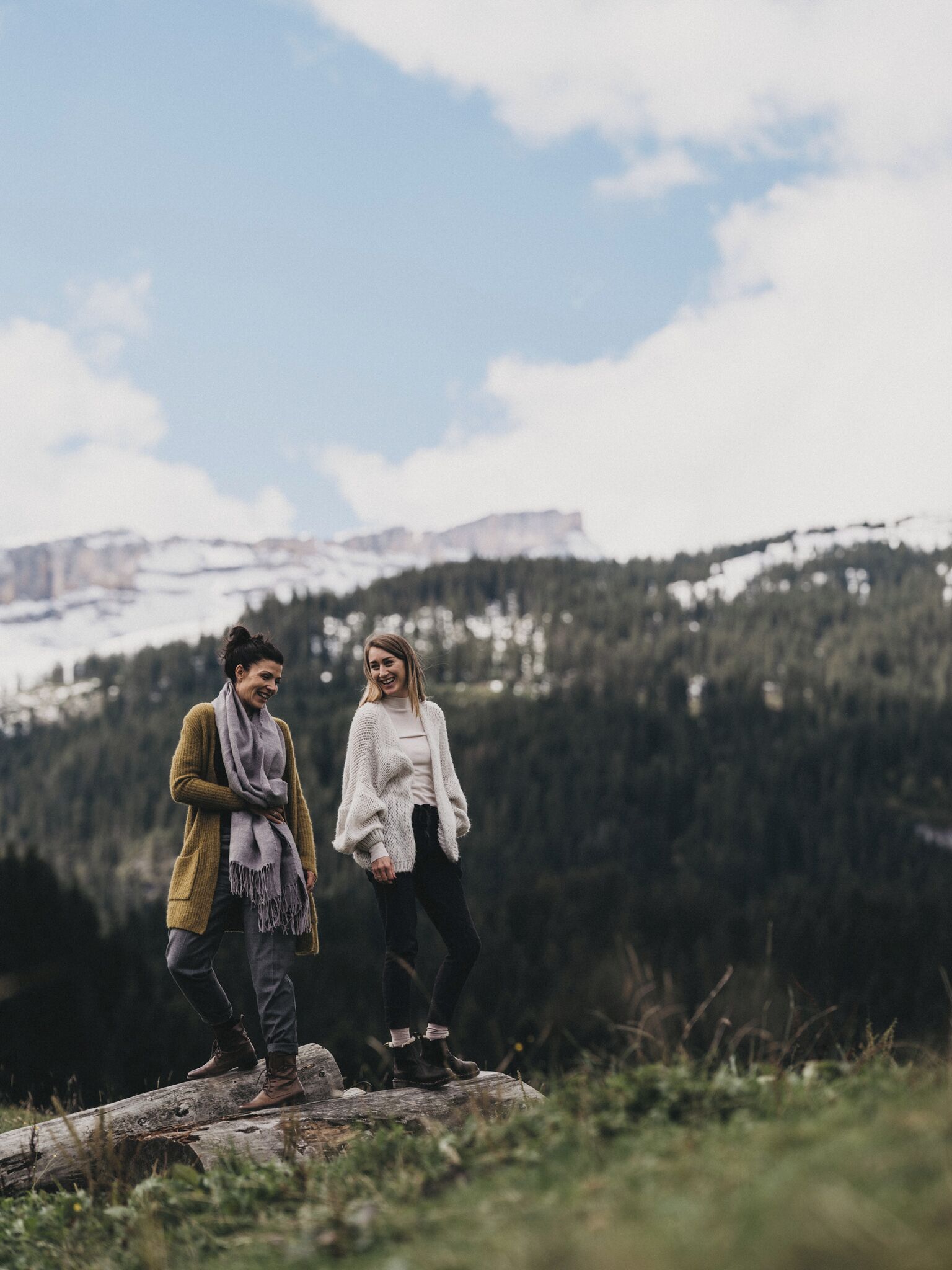 Dos personas de pie sobre un tronco en un entorno natural, con montañas al fondo.
