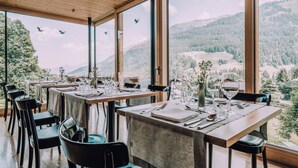 Comedor con mesas dispuestas para cuatro personas, amplias ventanas con vista a la montaña y techo de madera.