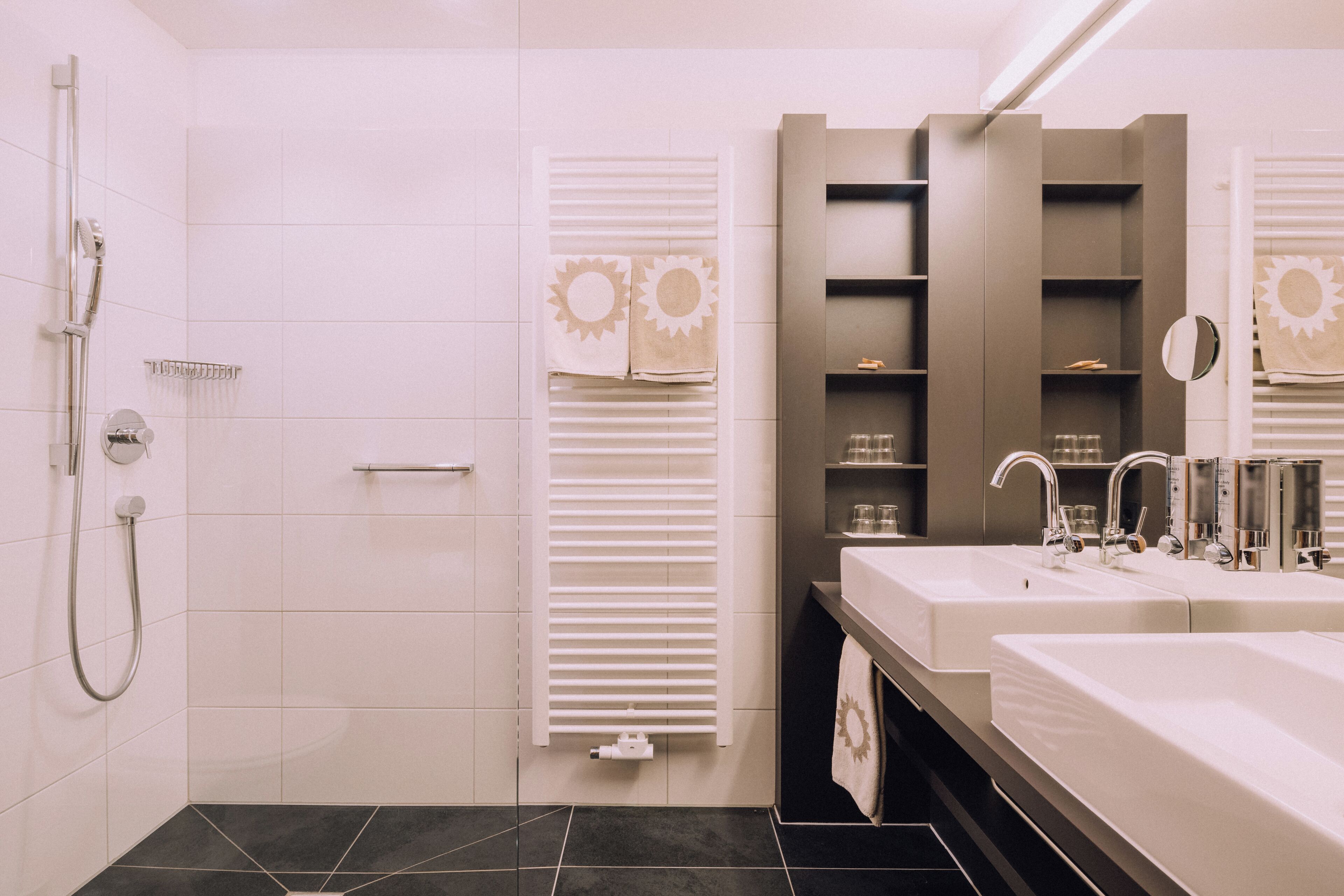 Un baño moderno con dos lavamanos, una ducha y un perchero con toallas con motivos de sol.