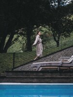 A person walking on a sloped pathway beside a pool with lounge chairs.