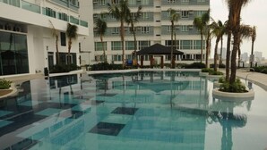 A swimming pool with a tiled pattern, surrounded by palm trees and a modern building with multiple floors. - The Beacon 1Br for 4, Tower 2, WiFi 300Mbps (Makati City)