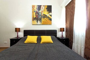 Quarto com uma cama grande, duas mesas de cabeceira, um quadro abstrato moderno e uma janela com cortinas.