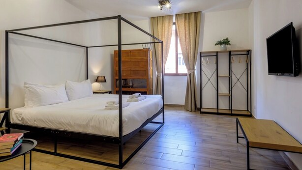 A four-poster bed with a wooden headboard, a bedside table with books, a wooden bench, a wardrobe with a mirror, and a TV on the wall. - Trastevere Home 14 Roma Centro, San Pietro, Gianicolo, American University. (Roma)
