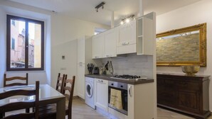 A compact kitchen with white cabinets, a built-in oven, a washing machine, and a dining area with wooden chairs. - Trastevere Home 14 Roma Centro, San Pietro, Gianicolo, American University. (Roma)