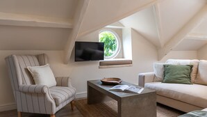 A cozy attic room with a TV, a window with a view of greenery, a striped armchair, a sofa, and a coffee table.