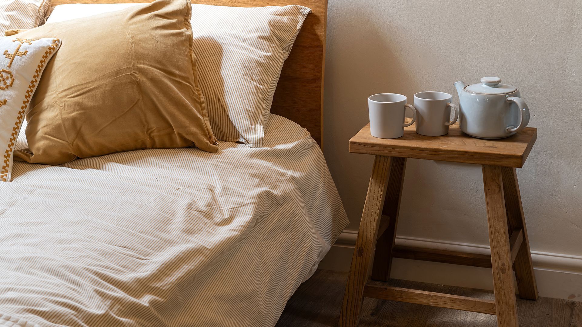 Een opgemaakt bed met een gestreept dekbed, een beige kussen en een houten nachtkastje met twee witte mokken en een theepot.