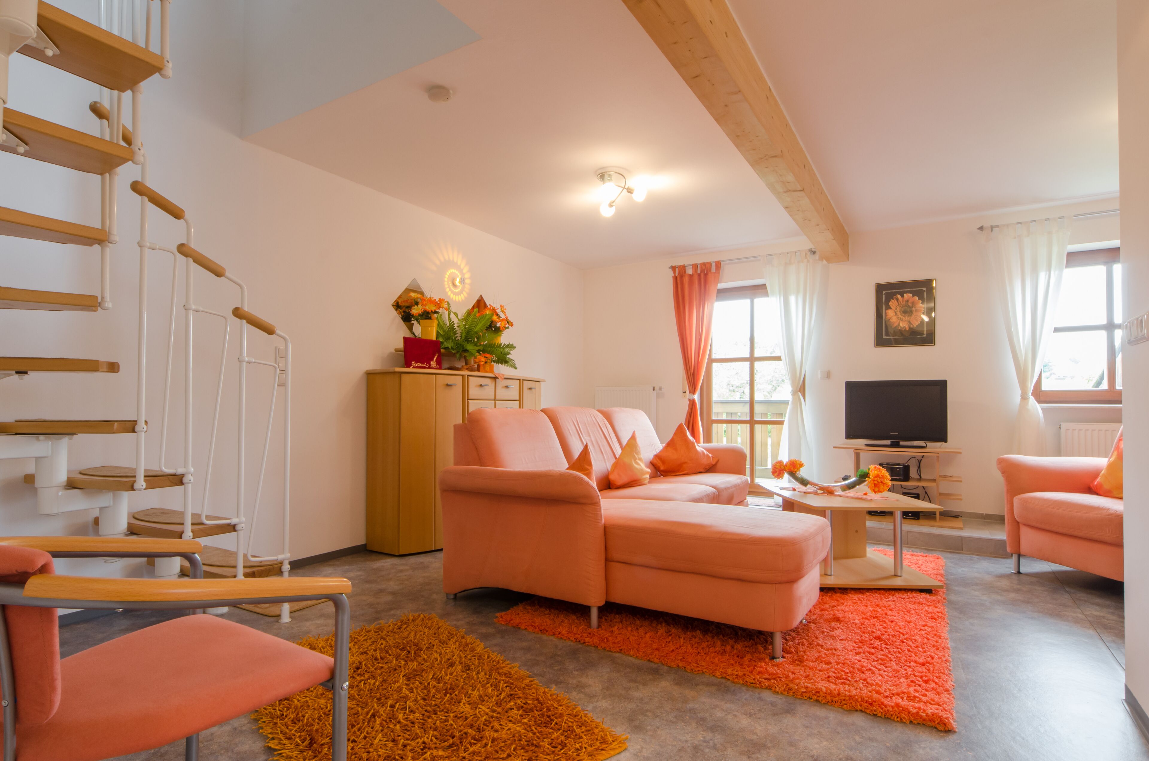 A modern living room with a pink sofa, orange rug, wooden staircase, and a TV.