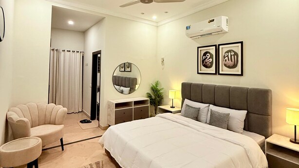 A modern bedroom with a large bed, a ceiling fan, an air conditioning unit, a round mirror, and framed artwork on the wall. - Designer & Spacious 2BHK Private Suite (Islamabad)