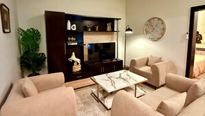 A living room with beige sofas, a coffee table, a TV stand with decorative items, and a clock on the wall. - Designer & Spacious 2BHK Private Suite (Islamabad)