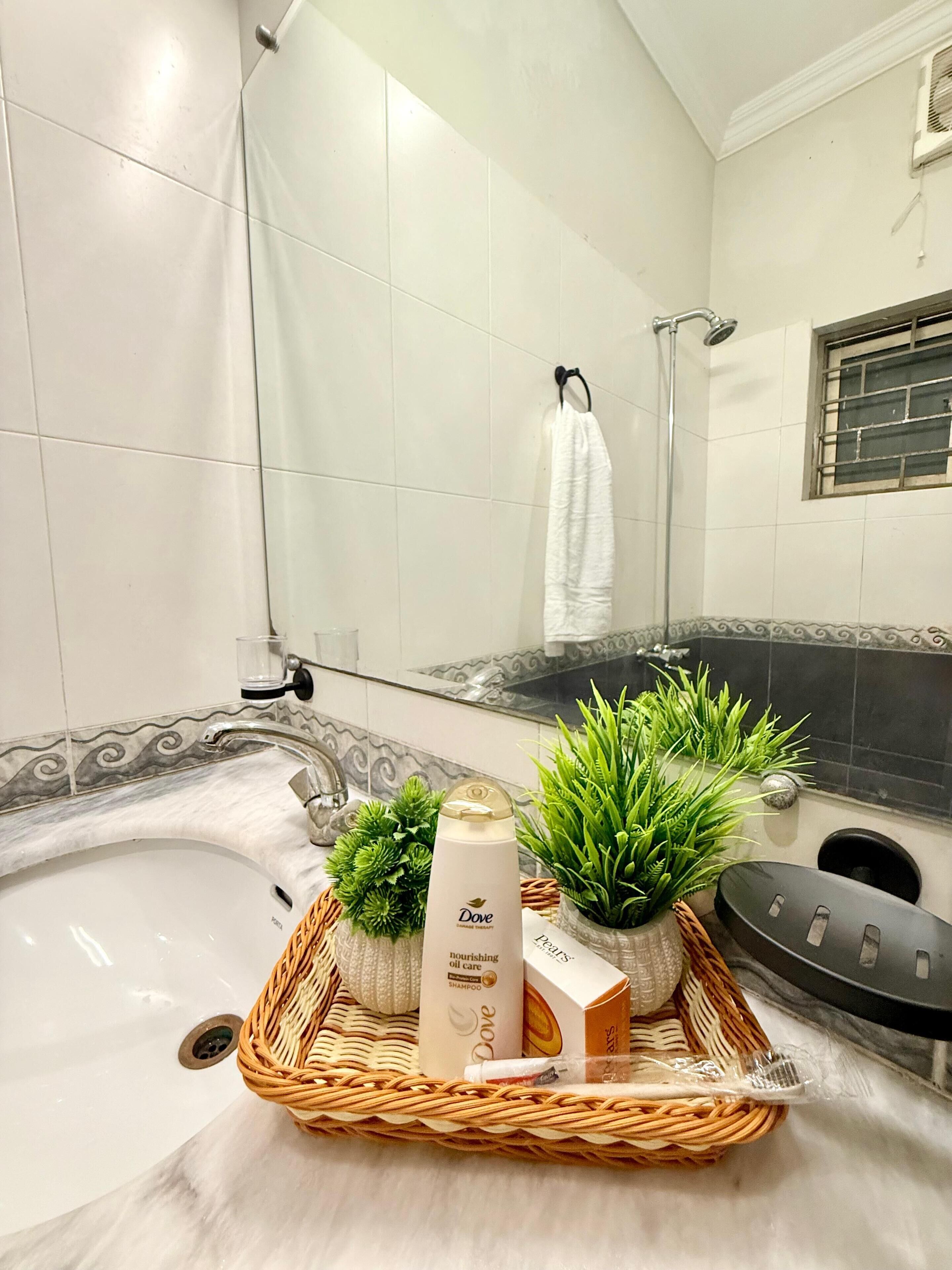 A bathroom with a bathtub, a shower, and a sink. There are plants, a bottle of Dove shampoo, and a basket with toiletries.