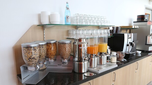 A breakfast bar with dispensers of granola, cereal, and orange juice, a coffee machine, and a water dispenser. - Ibis Budget Augsburg City (Augsburg)