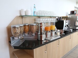 A breakfast bar with dispensers for cereals, milk, and orange juice, and a coffee machine. - Ibis Budget Augsburg City (Augsburg)