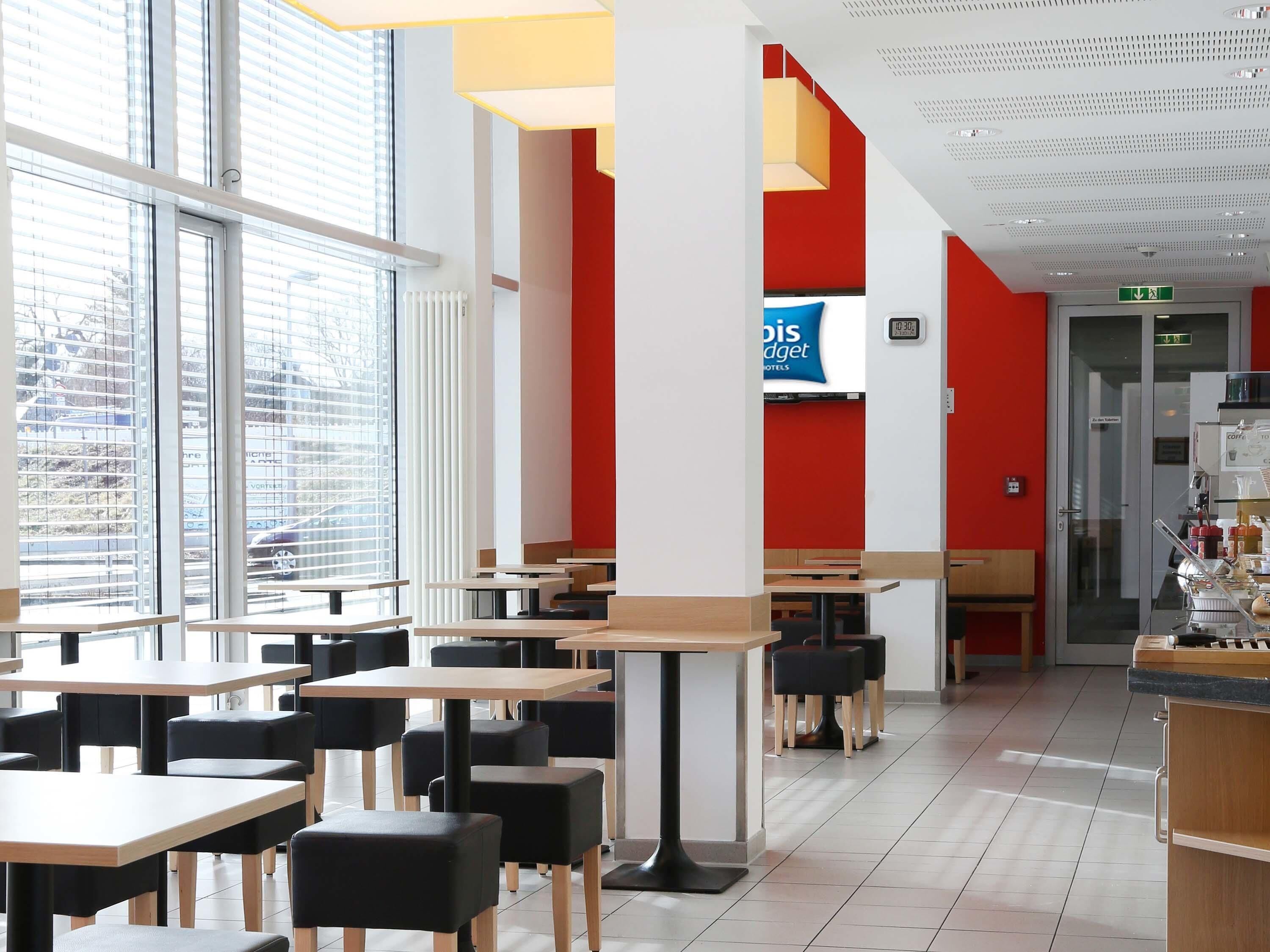 Un restaurant moderne avec des tables et des chaises, de grandes fenêtres et un mur d’accent de couleur rouge.
