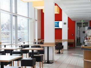 Ein modernes Restaurant mit Tischen, Stühlen, großen Fenstern und einer roten Akzentwand.